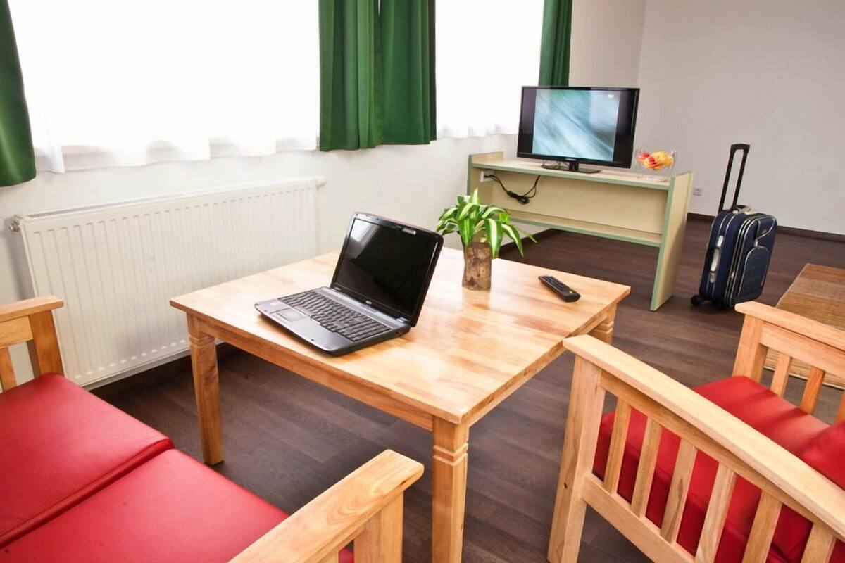 A room with a wooden table, a laptop, a plant, a television, and a suitcase.