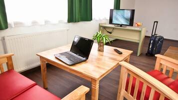 Una habitación con una mesa de madera, una computadora portátil, una planta, un televisor y una maleta.