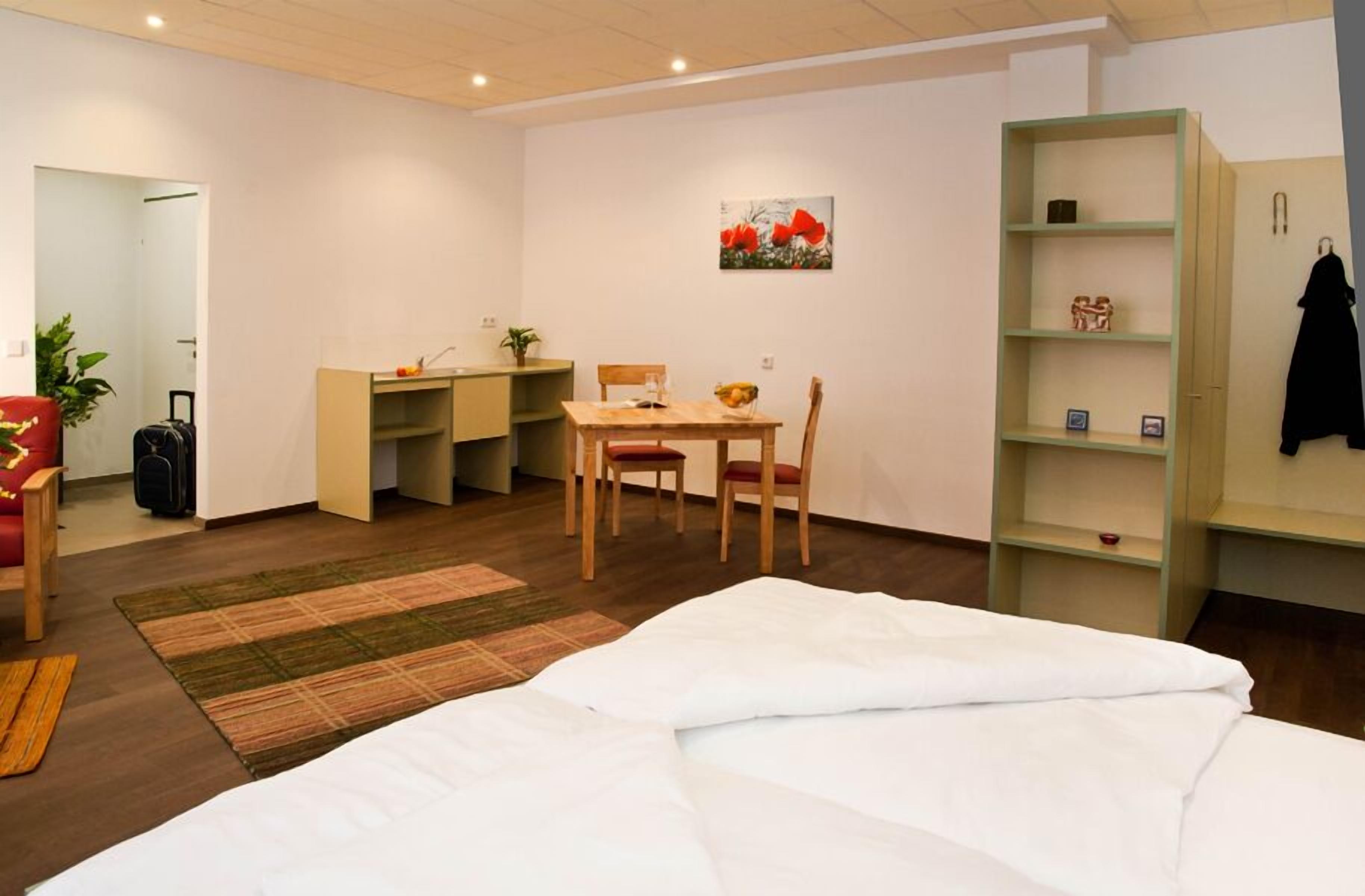 A hotel room with a bed, a desk, a chair, a small table, a bookshelf, and a painting on the wall.
