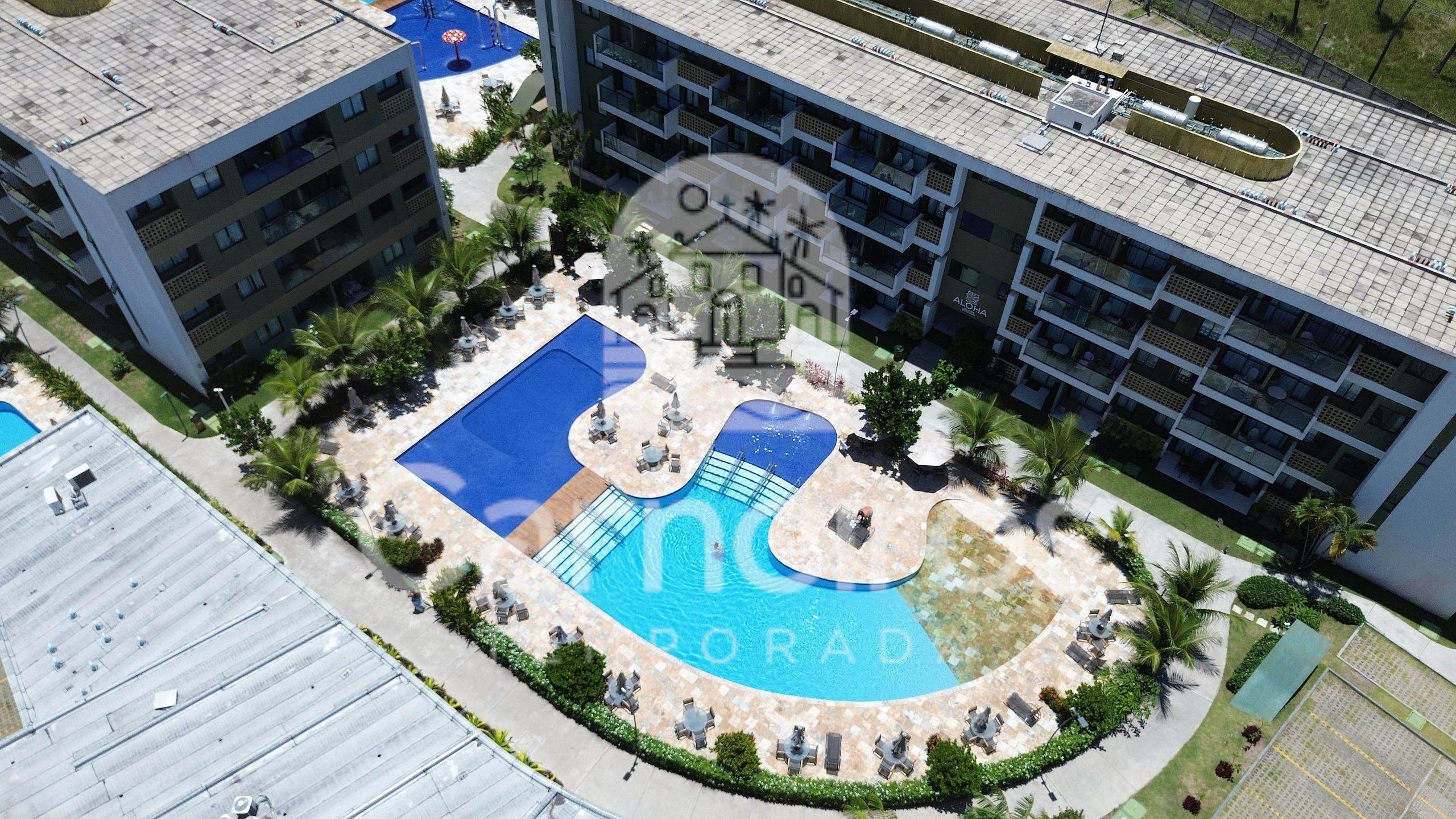 A hotel with a central swimming pool surrounded by lounge areas and palm trees.