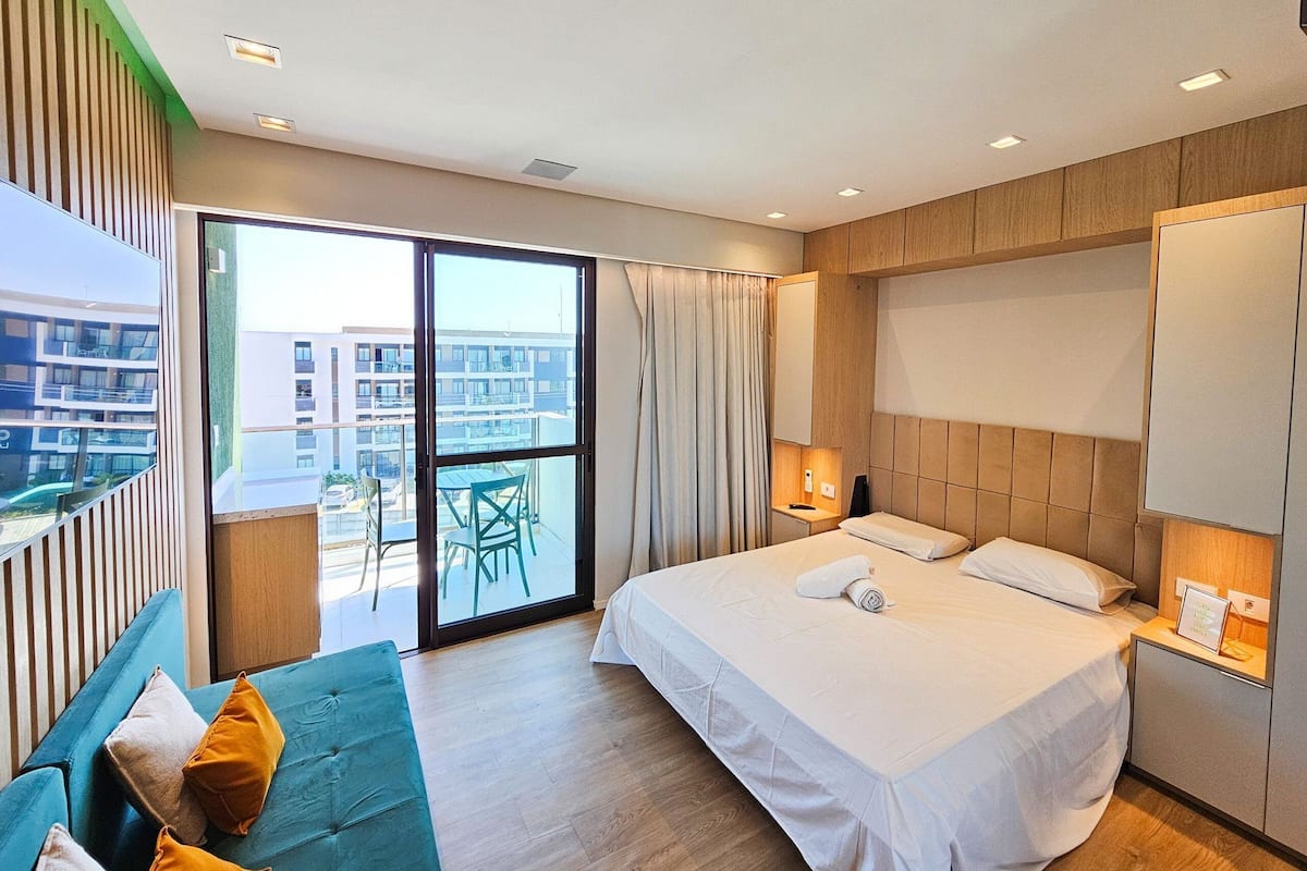 Quarto de hotel moderno com uma cama grande, um sofá, uma varanda com vista para edifícios e uma porta de correr de vidro.