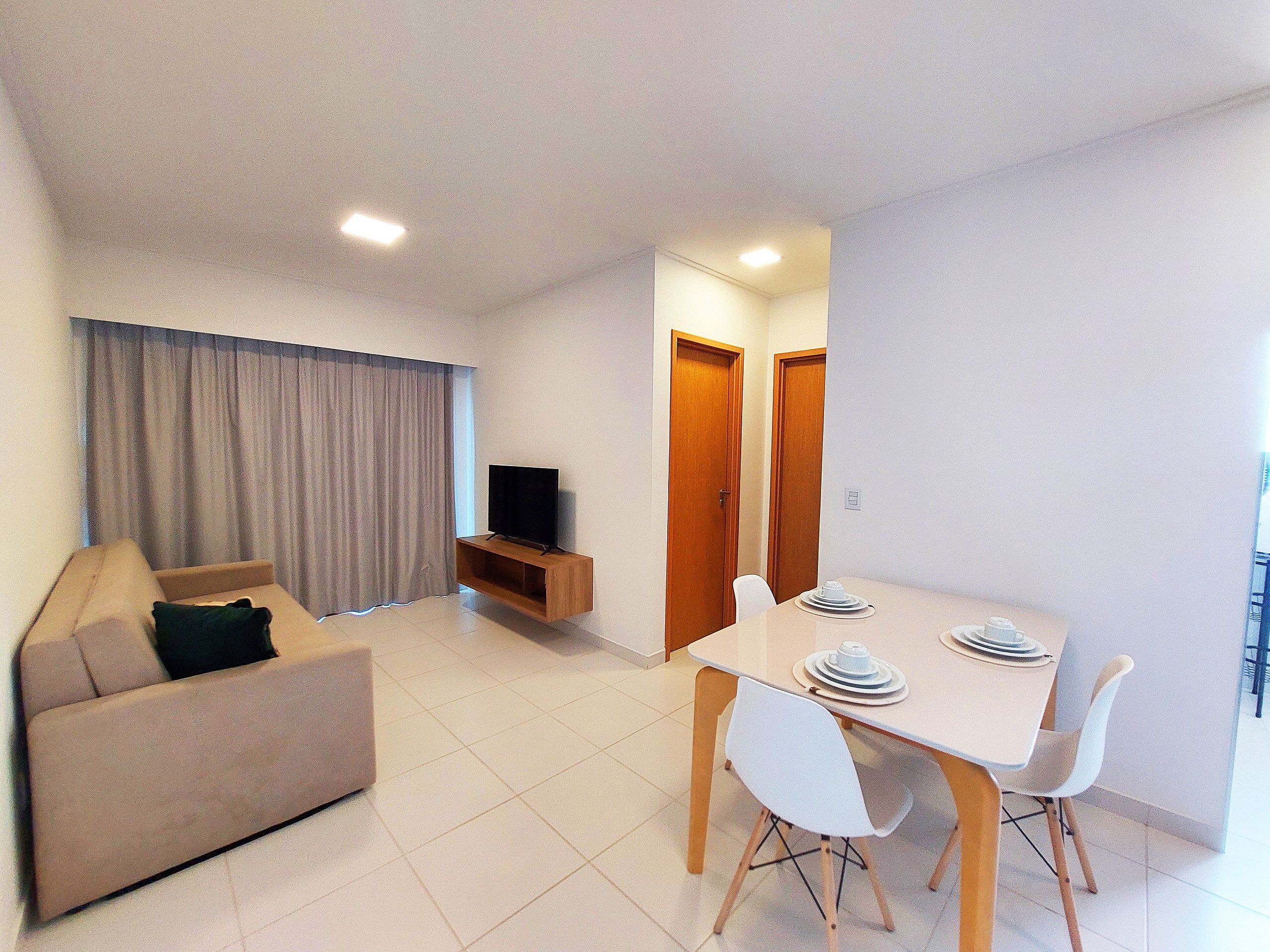 A living room with a sofa, a TV on a wooden stand, a dining table with chairs, and a kitchen area with cabinets.