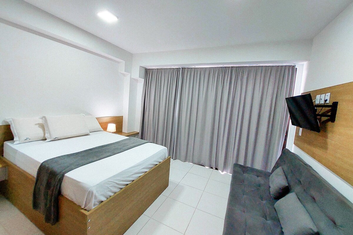 A hotel room with a bed, a bedside table with a lamp, a TV mounted on the wall, and a sofa.