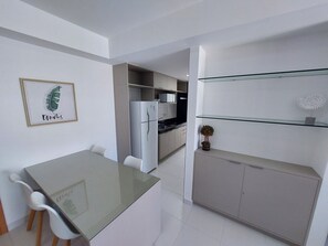 A modern kitchen with a refrigerator, a dining table with chairs, and a glass shelf unit. - Mana Beach Experience- Porto de Galinhas (Ipojuca)