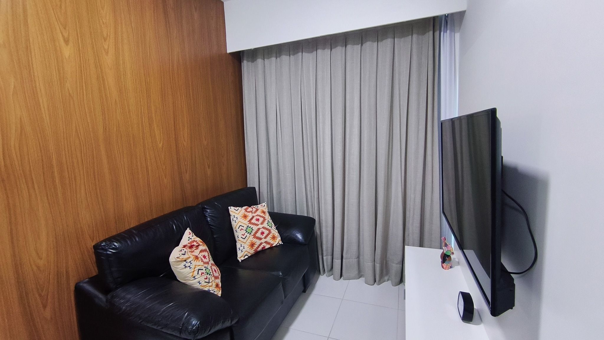 Een woonkamer met een zwarte leren bank, twee gestreepte kussens, een grijs gordijn en een wandgemonteerde flatscreen-tv.