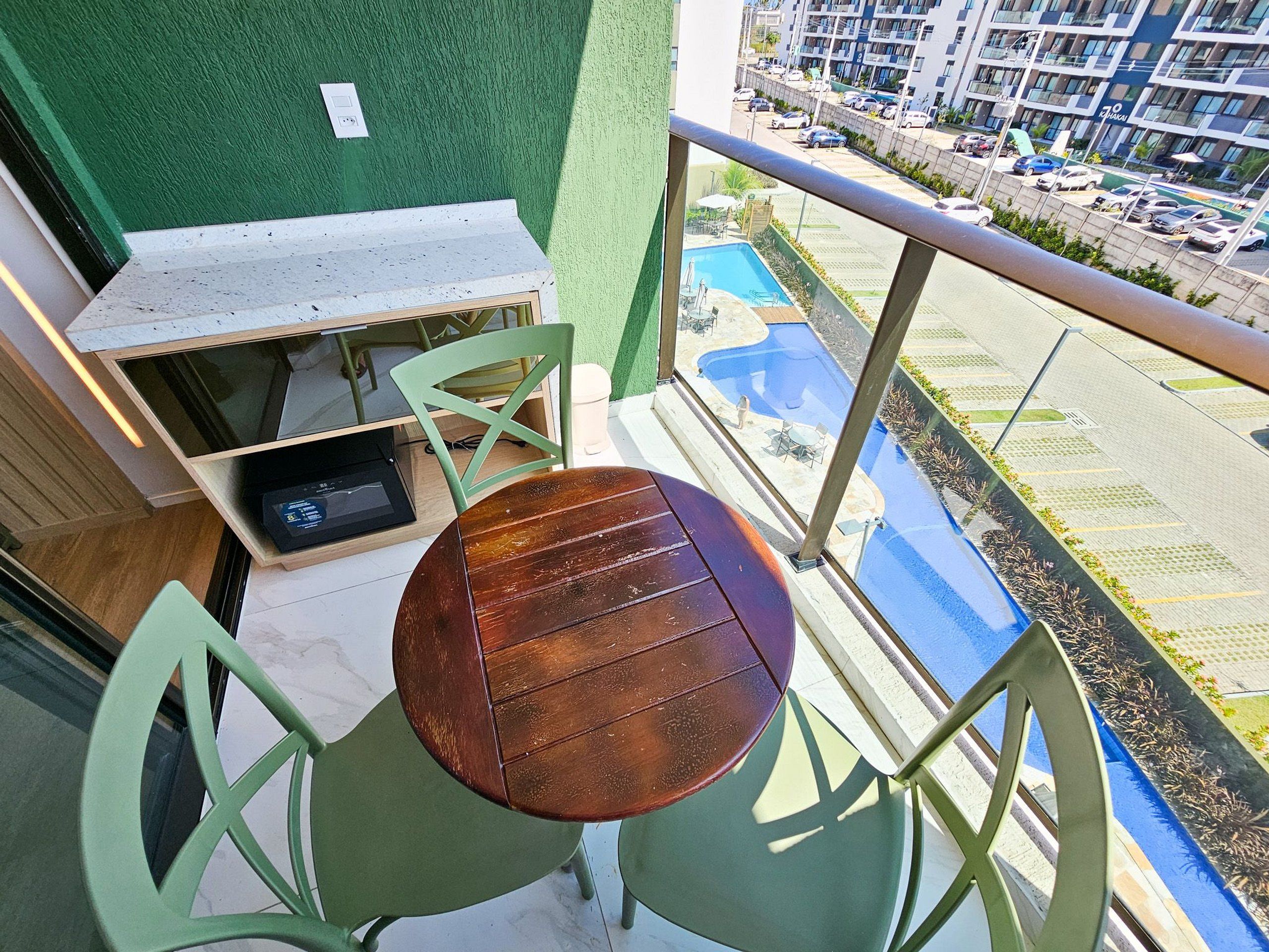 Un balcon avec une table et des chaises en bois, un petit bureau et une vue sur une piscine et des immeubles.