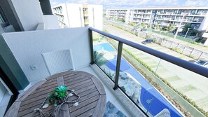 A balcony with a wooden table, a potted plant, and a view of a swimming pool and residential buildings. - Mana Beach Experience- Porto de Galinhas (Ipojuca)