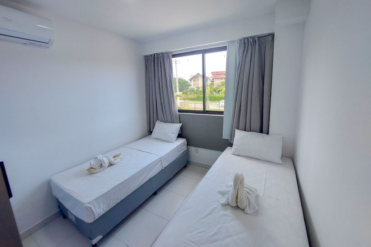 A small hotel room with two beds, a window with curtains, a visible air conditioning unit, and a view of buildings outside.