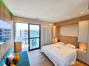A modern hotel room with a large bed, a sofa, a balcony with a view of buildings, and a sliding glass door. - Mana Beach Experience- Porto de Galinhas (Ipojuca)