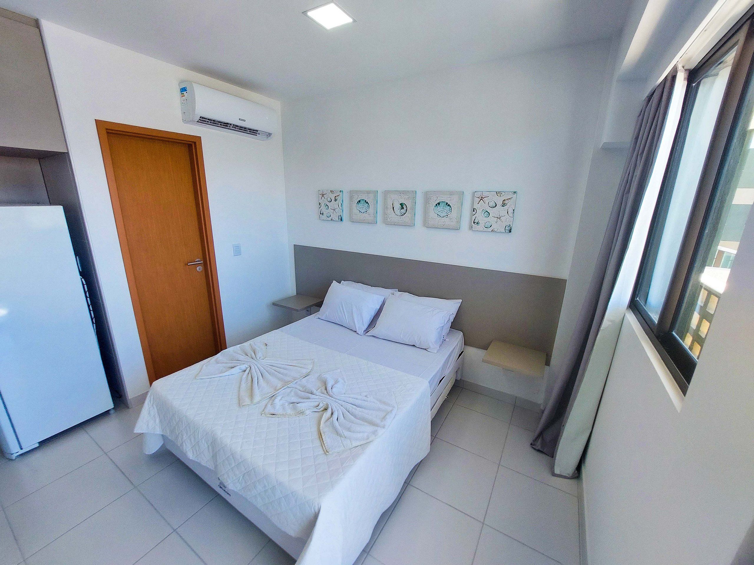 A hotel room with a bed, a small bedside table, a wall-mounted air conditioner, and a window with curtains.