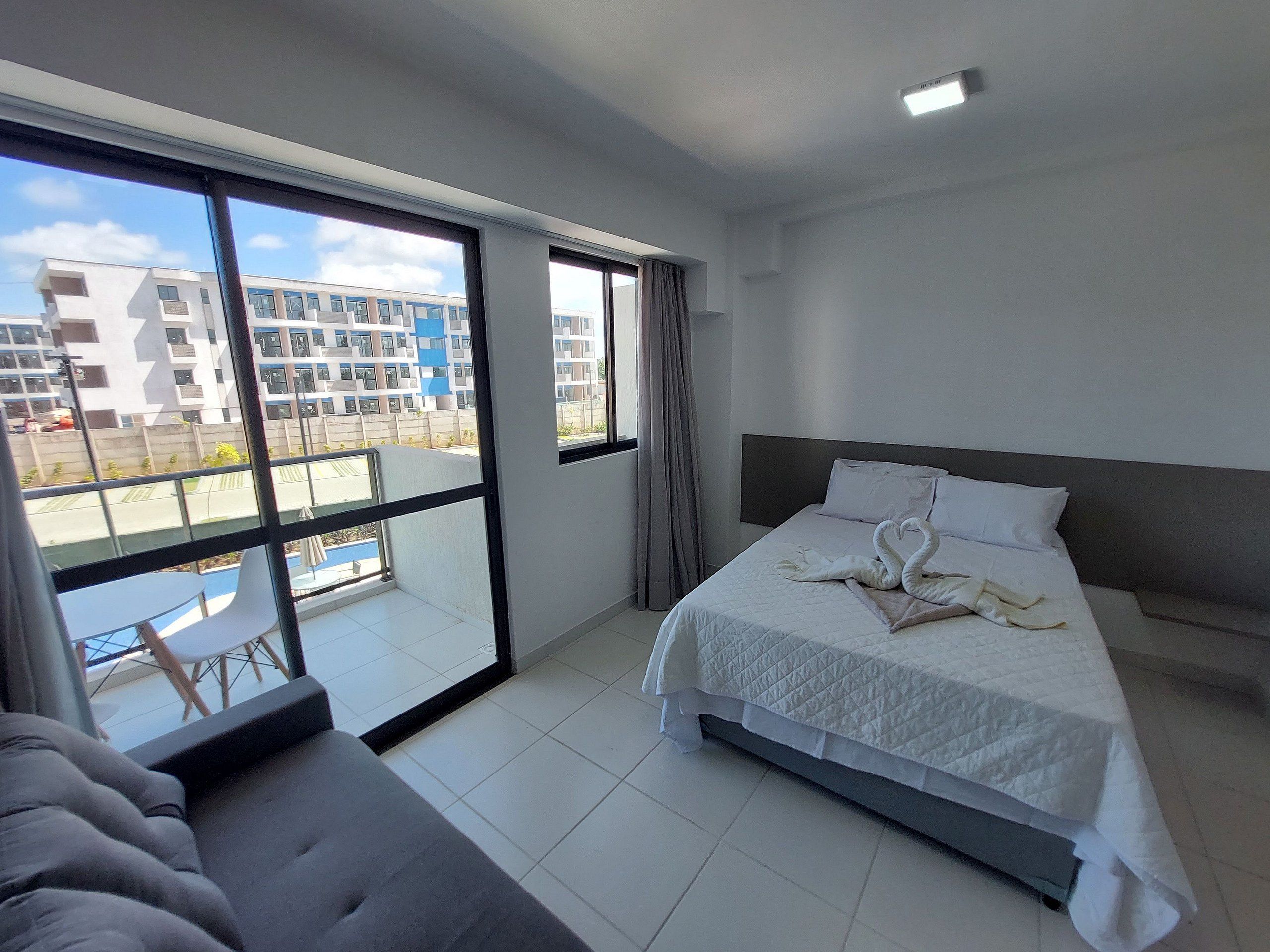 Habitación de hotel con cama, sofá y ventanas amplias con vistas a edificios.