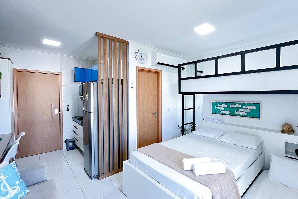 Quarto de hotel moderno com cama, escrivaninha, guarda-roupa e prateleira com objetos decorativos.