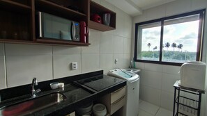 A kitchen with a microwave, stove, sink, and window with a view of palm trees. - Mana Beach Experience- Porto de Galinhas (Ipojuca)