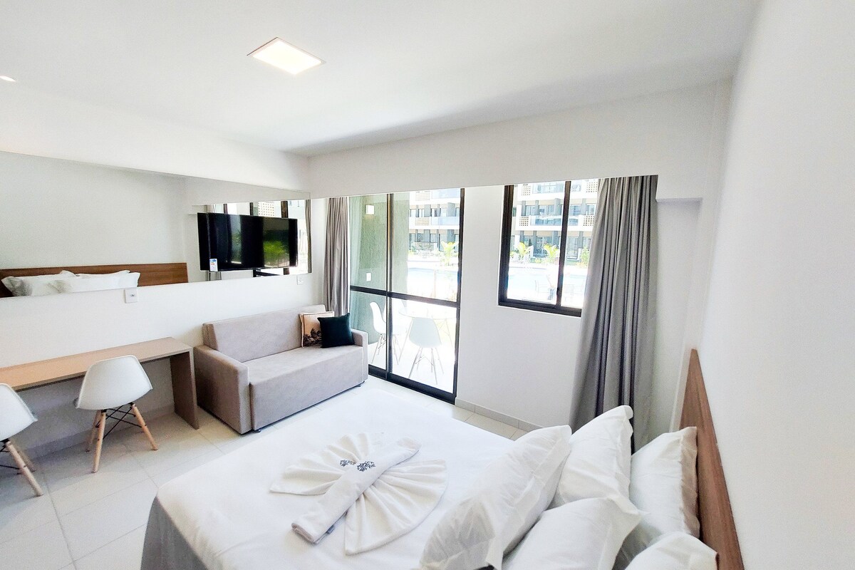 A hotel room with a bed, a sofa, a desk, a TV, and large windows.
