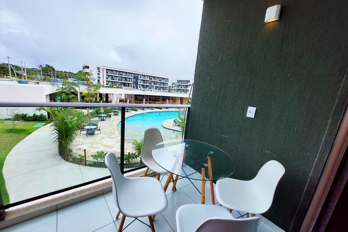 Uma varanda com mesa e cadeiras de vidro, com vista para uma piscina e o prédio do hotel.