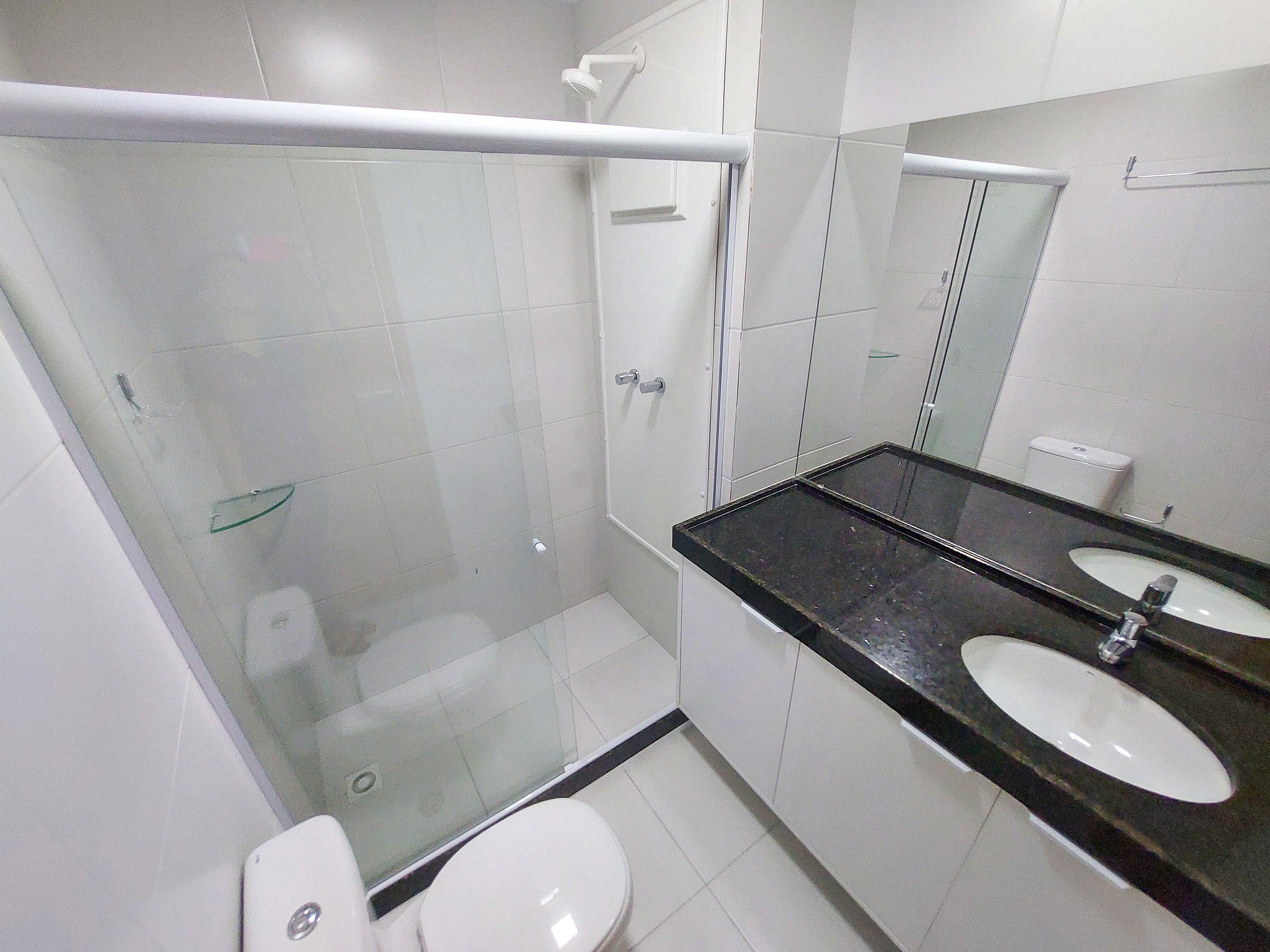 Un baño con mampara de vidrio, inodoro blanco y encimera negra con dos lavamanos.