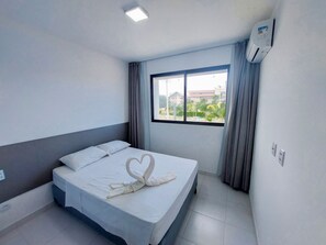 A hotel room with a bed, a window with curtains, an air conditioning unit, and a view of buildings and greenery outside. - Mana Beach Experience- Porto de Galinhas (Ipojuca)