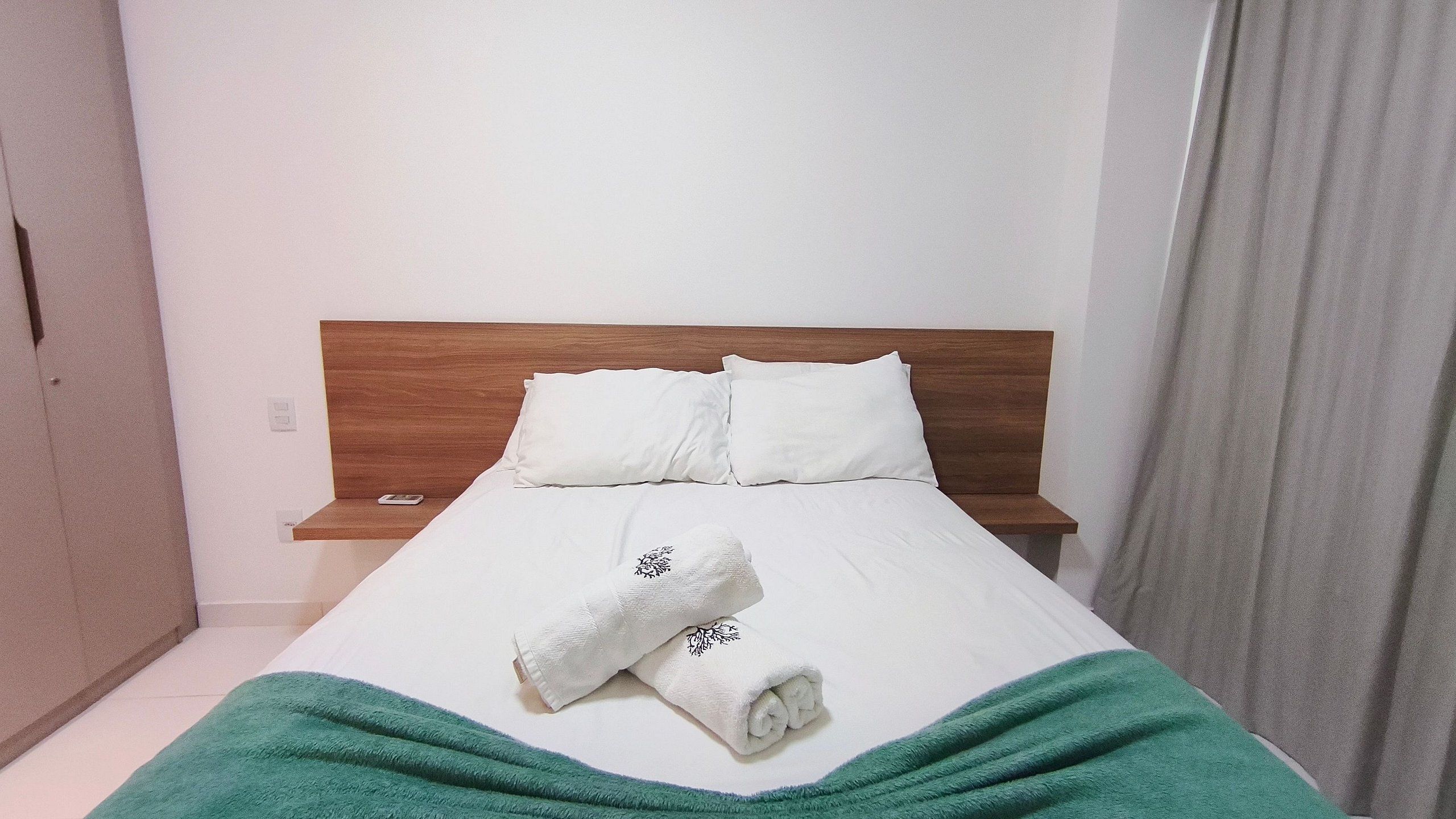 A hotel room with a wooden headboard, white bedding, a green blanket, and a folded towel on the bed.