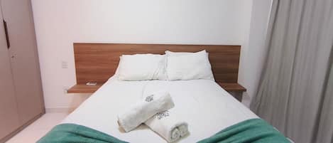 A hotel room with a wooden headboard, white bedding, a green blanket, and a folded towel on the bed.
