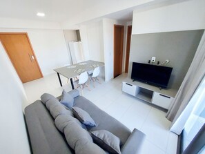 A modern living room with a grey sofa, a dining table with chairs, a flat-screen TV, and a wooden door. - Mana Beach Experience- Porto de Galinhas (Ipojuca)