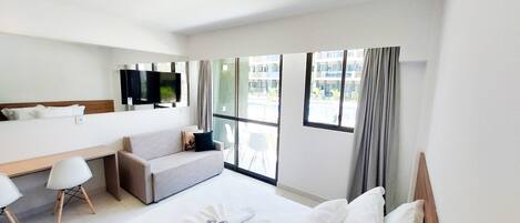A hotel room with a bed, a sofa, a desk, a TV, and large windows.