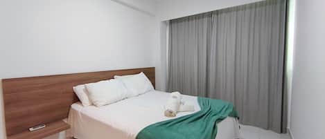 A hotel room with a wooden headboard, a bed with white bedding, a green throw blanket, and a window with grey curtains.