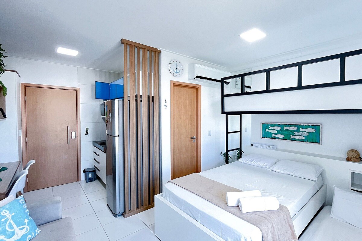 A modern hotel room with a bed, a desk, a wardrobe, and a wall-mounted shelf with decorative items.