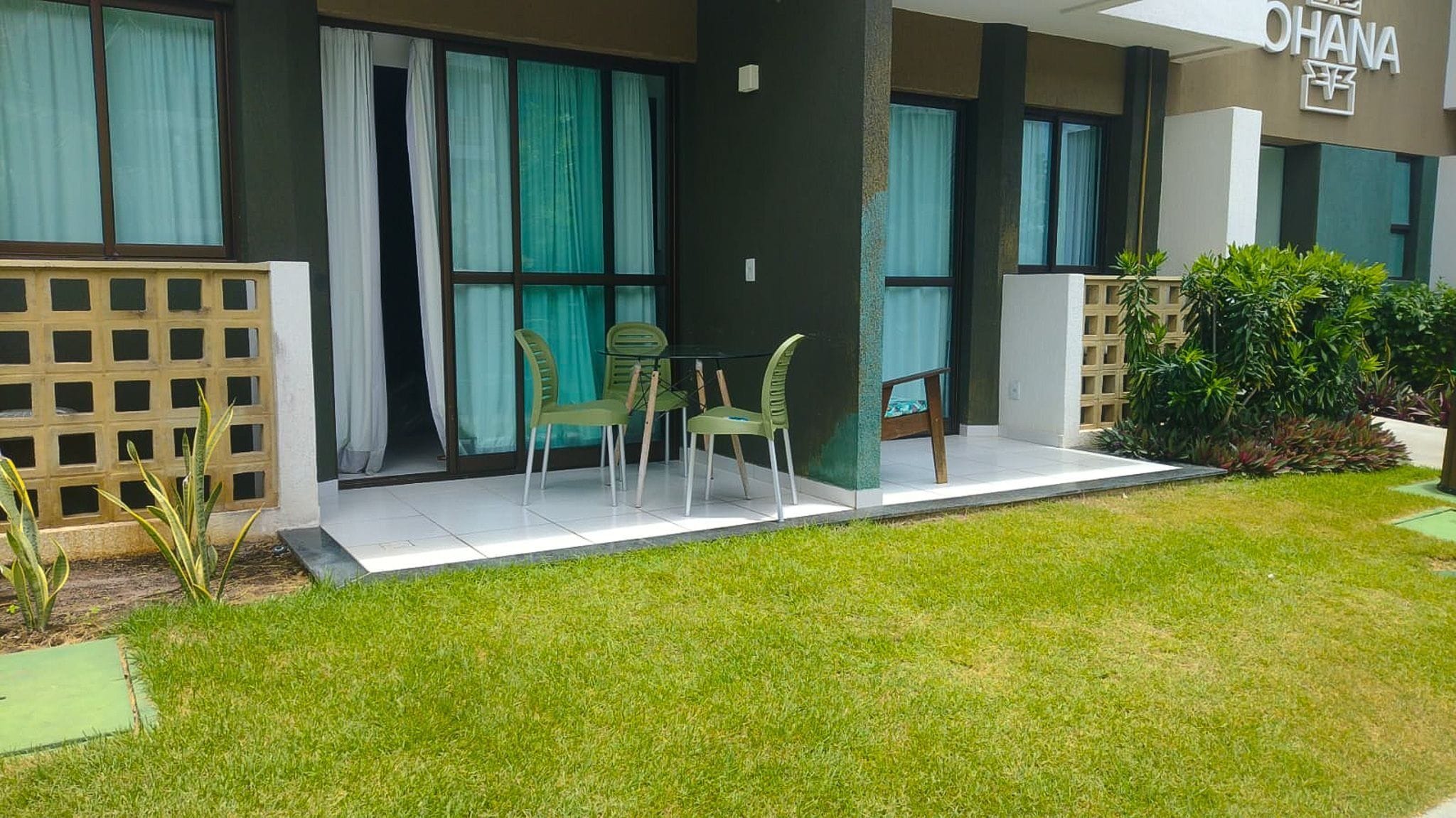 Exterior moderno de un hotel con un área de estar al aire libre cubierta, un césped cuidado y un cartel con el nombre del hotel.