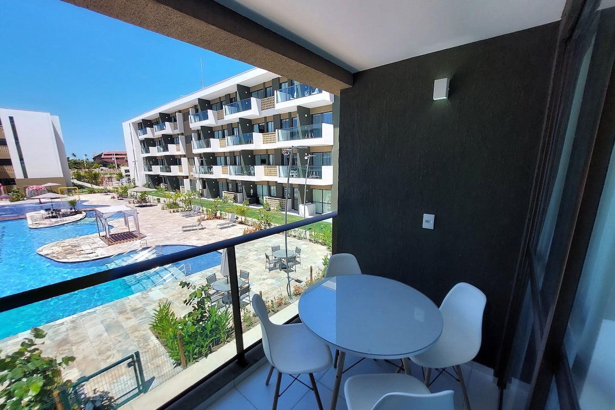 Varanda com vista para a piscina, cadeiras brancas e um edifício moderno ao fundo.