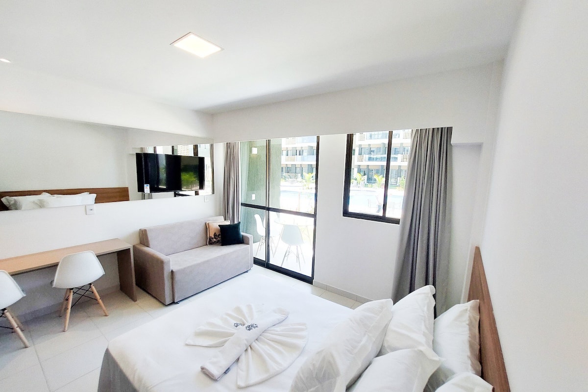 Quarto de hotel com cama, sofá, escrivaninha, televisão e grandes janelas.
