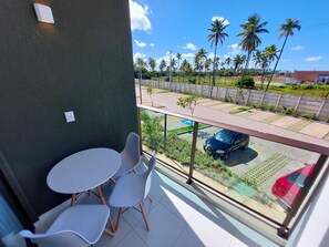 A balcony with a table and chairs, a view of a road with palm trees, and a red car parked outside. - Mana Beach Experience- Porto de Galinhas (Ipojuca)