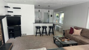 Uma sala de estar moderna com um sofá, uma mesa de centro de vidro e uma televisão. Há uma área de cozinha com um banco de bar e uma mesa de jantar.