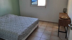 A bedroom with a bed, a small window, a wooden side table, and a radiator. - Comfortable T3 with terrace 5 minutes walk from Saint-Leu beach (Saint-Leu)