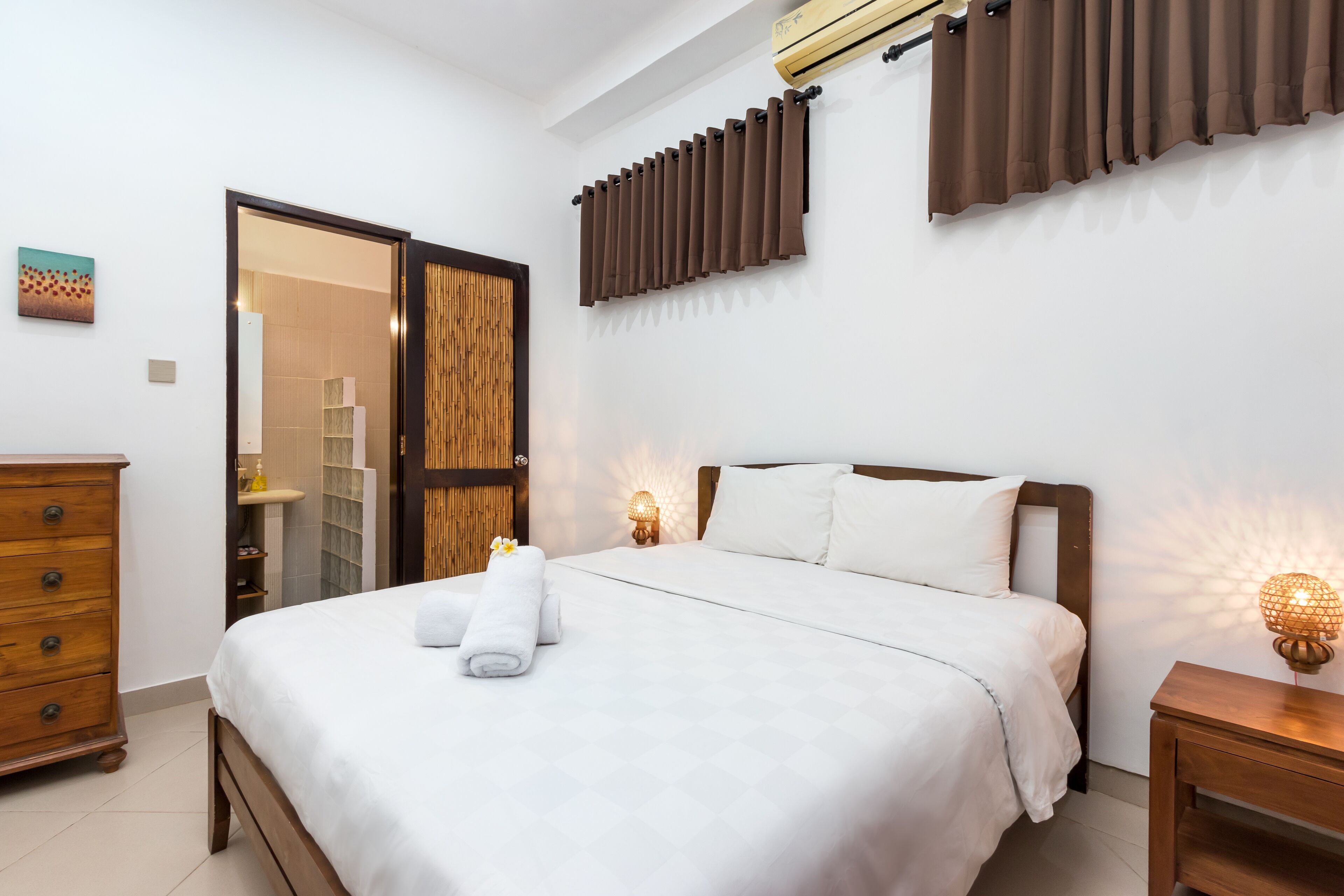 Une chambre à coucher bien rangée, avec un lit, des tables de chevet, une commode et une salle de bain visible à travers une porte ouverte.