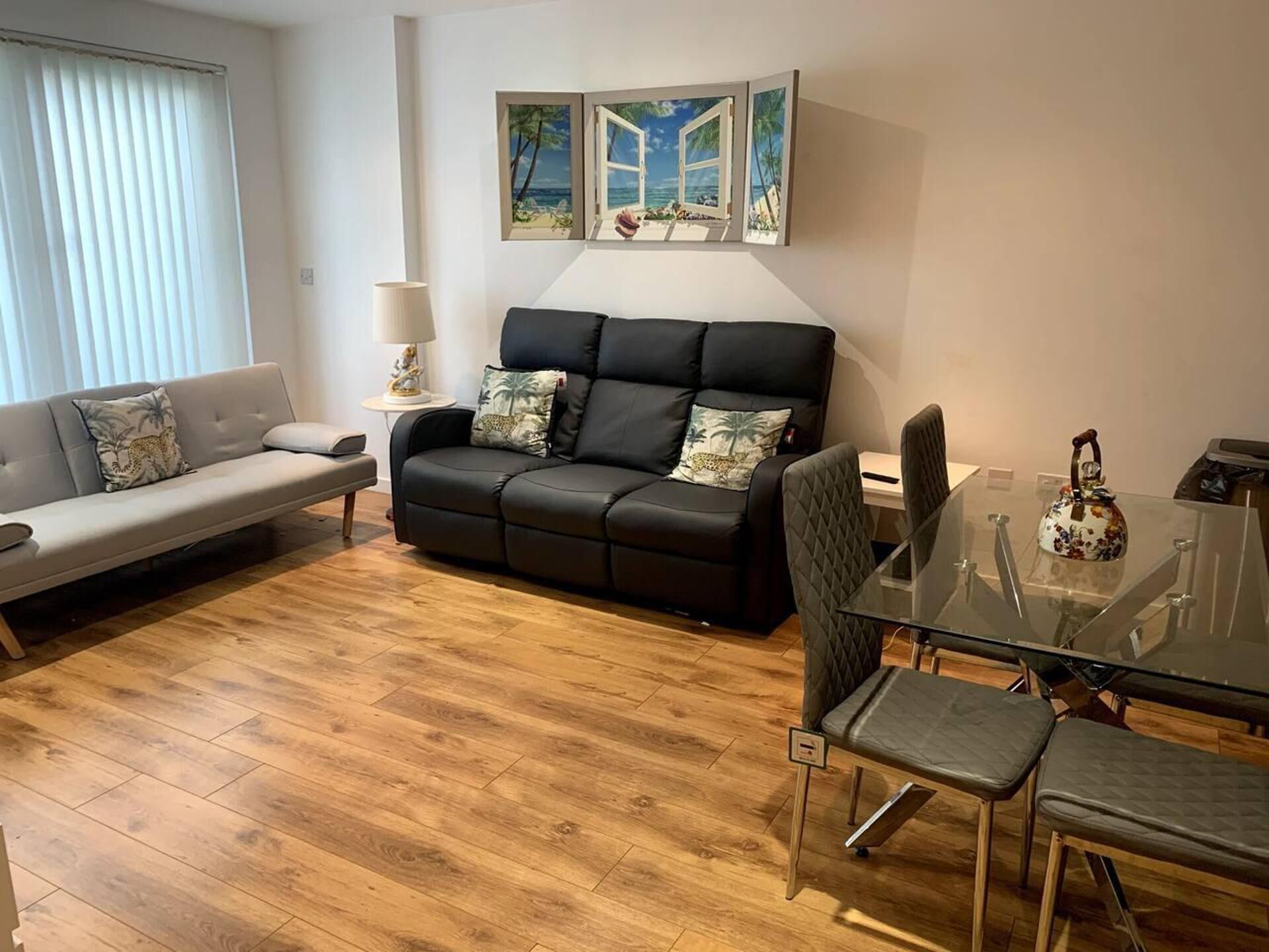 A modern living room with a black sofa, a grey sofa, a glass dining table with chairs, and a painting of a beach scene above the sofa.