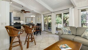 A spacious living area with a kitchen, dining table, and a balcony with a view of greenery.