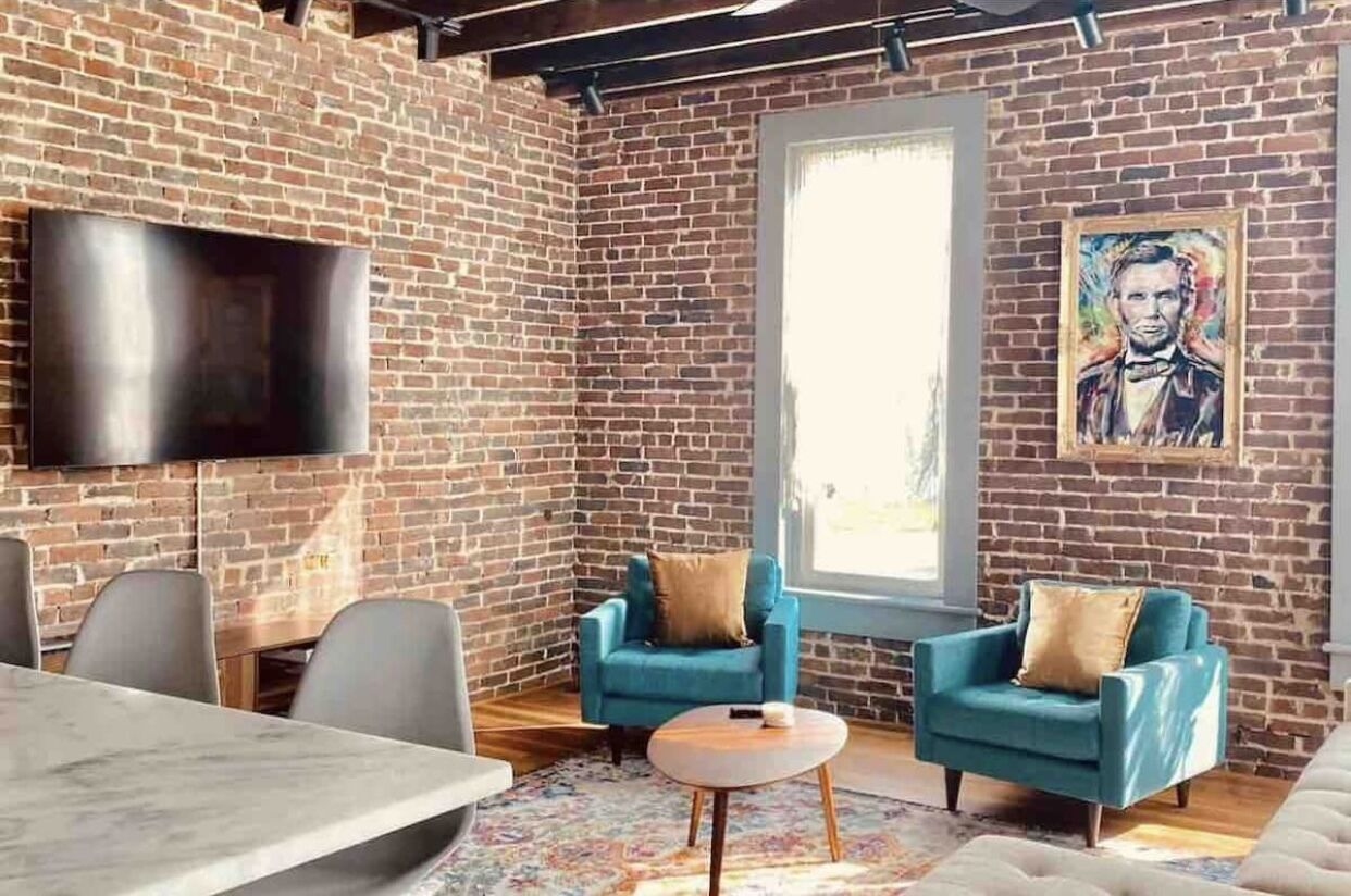 A modern interior with exposed brick walls, a flat-screen TV, two blue armchairs, a round coffee table, and a large window.
