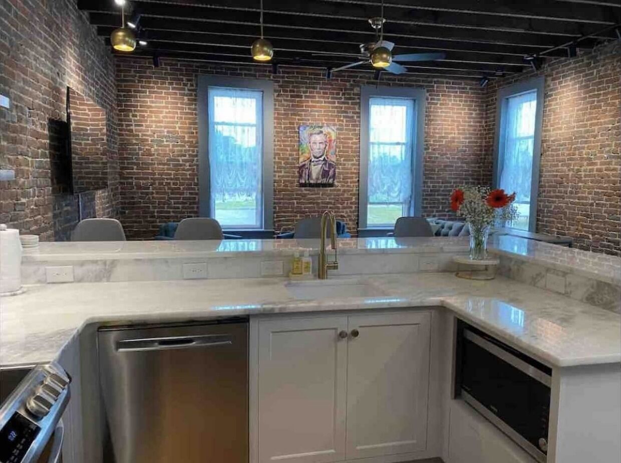 Eine Küche mit einer großen Kochinsel, einem Geschirrspüler und einem Spülbecken. Es gibt drei Fenster mit Vorhängen, einen Deckenventilator und eine Backsteinwand.