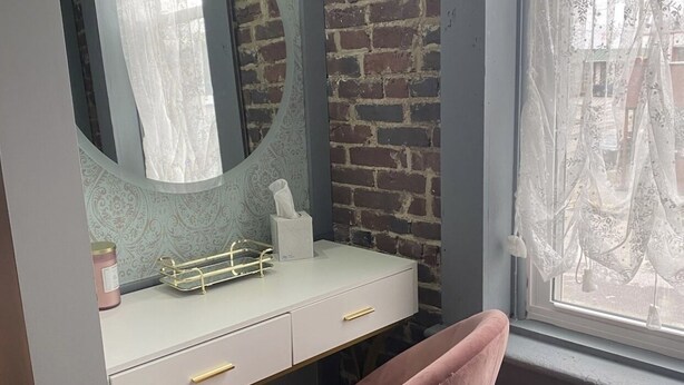 Une pièce avec une coiffeuse blanche, une chaise rose, un miroir rond et un mur en briques.