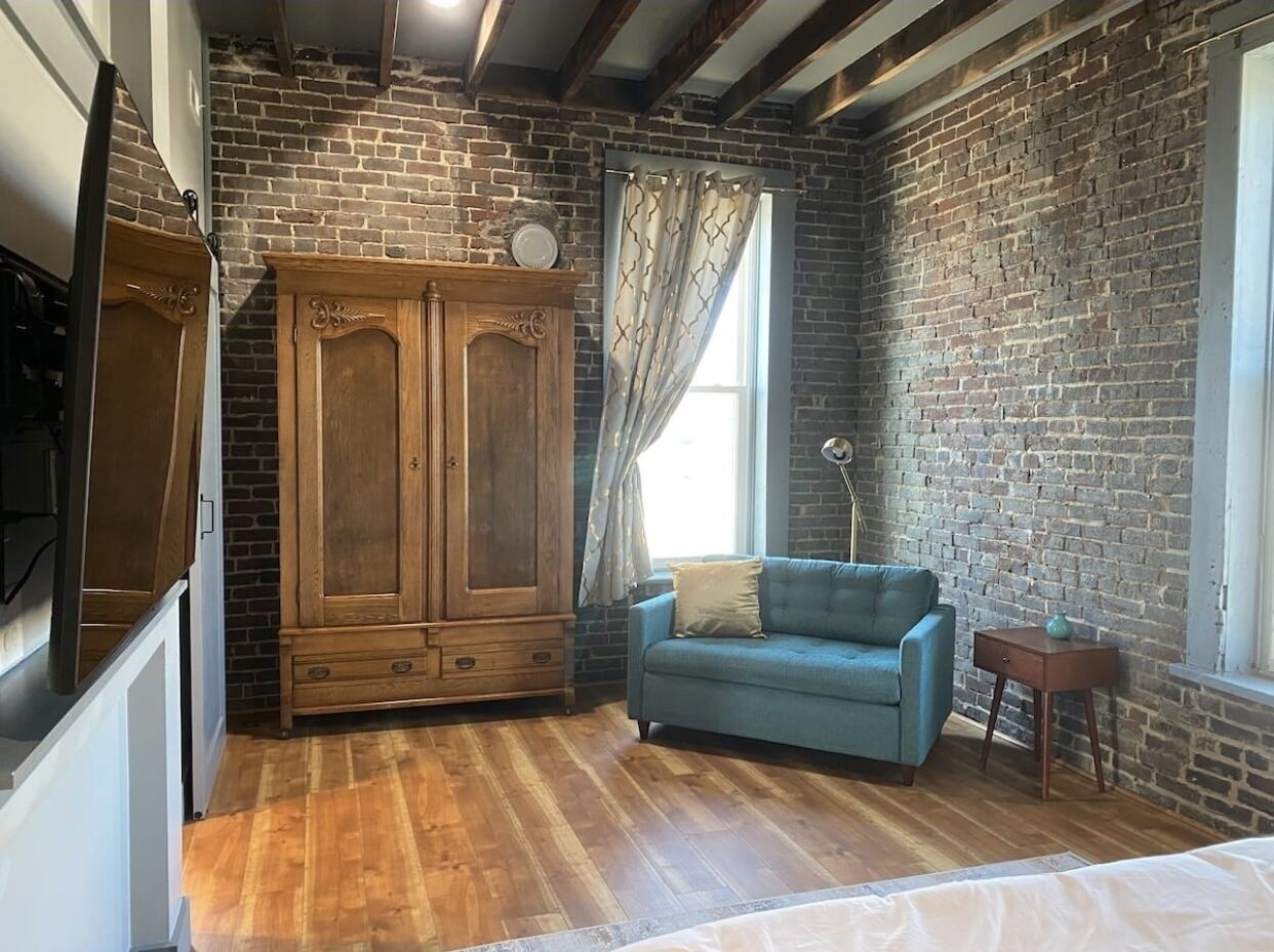 Ein Zimmer mit freiliegenden Backsteinwänden, einem Holzschrank, einem blauen Sofa und einem kleinen Tisch mit einer Lampe.