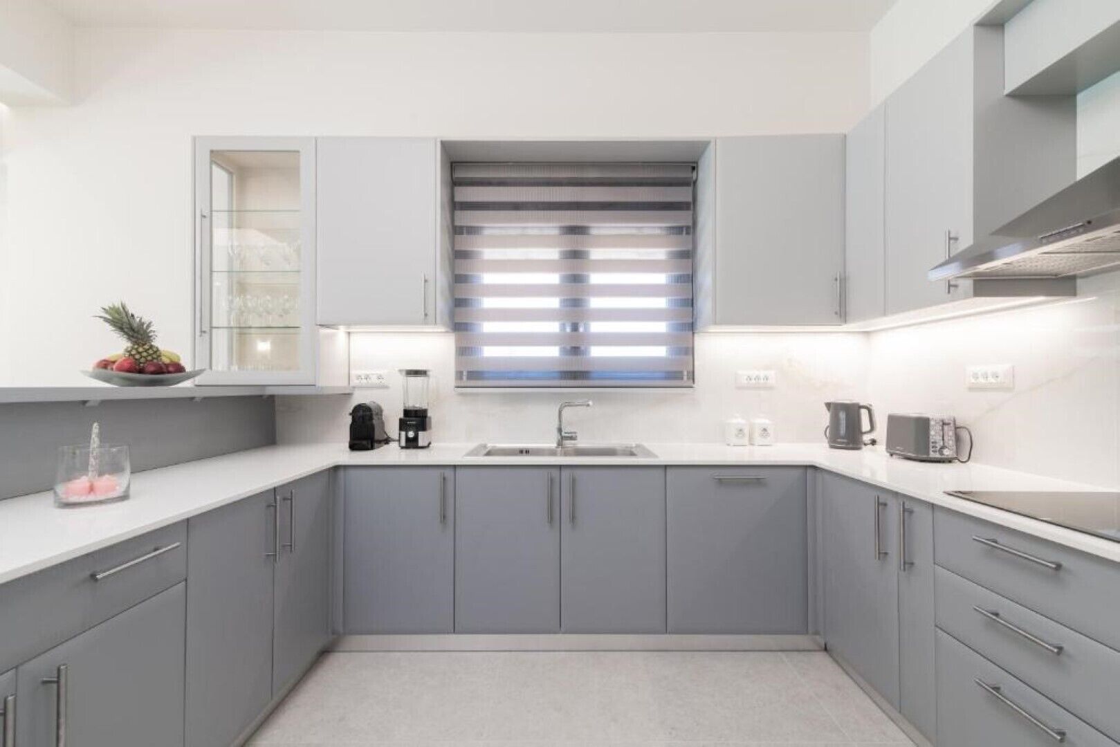 A modern kitchen with grey cabinets, a white countertop, a built-in window with blinds, and a coffee maker.