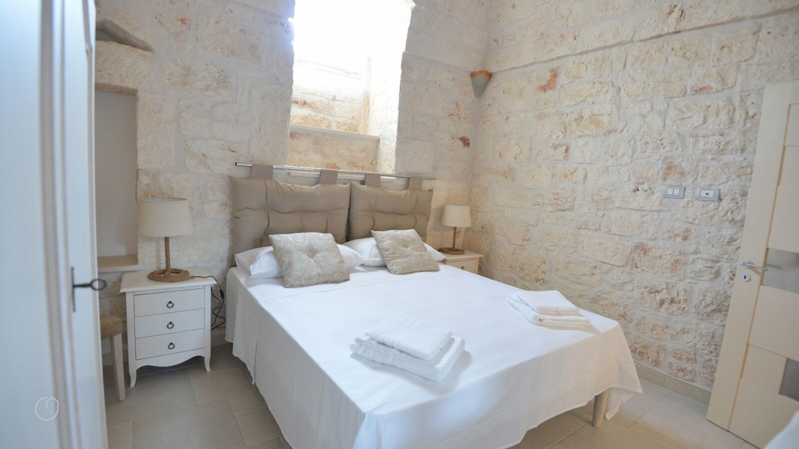 A bedroom with a stone wall, a bed with white linens, two bedside tables with lamps, and a door.