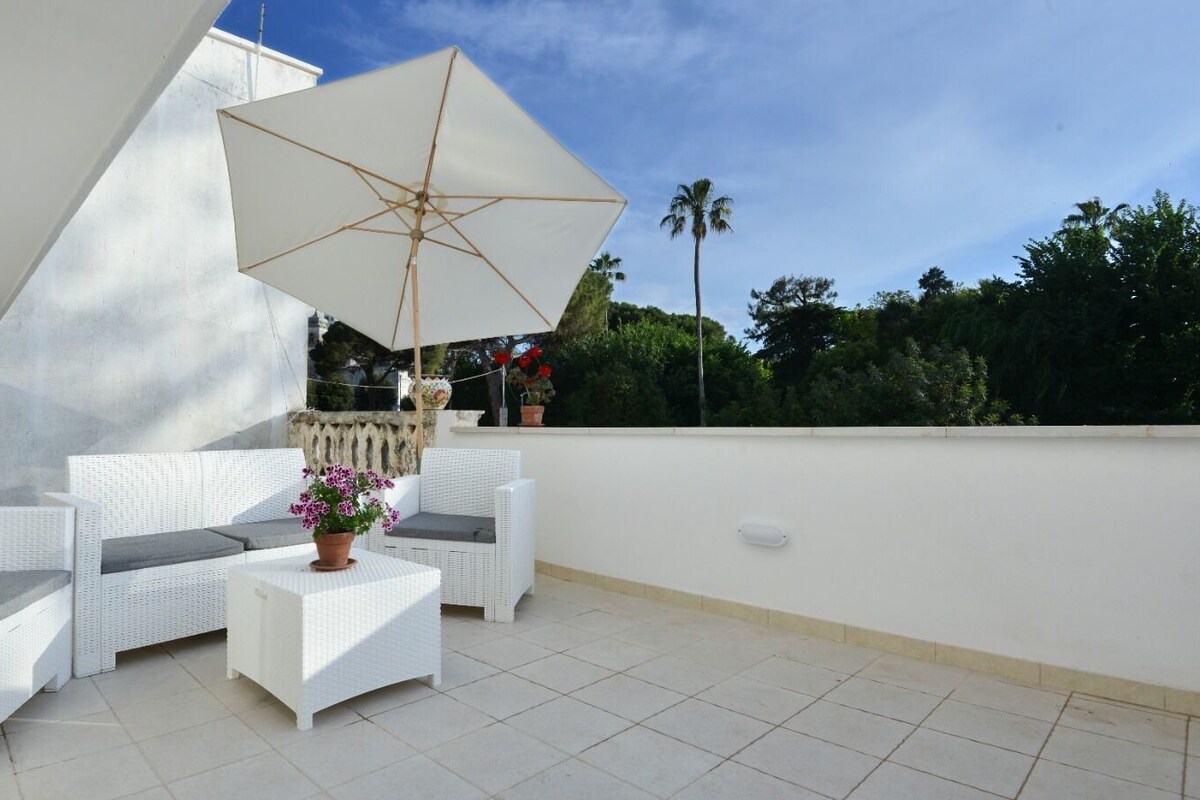 Un patio con mobiliario de mimbre blanco, una mesa con una planta en maceta y un paraguas grande.