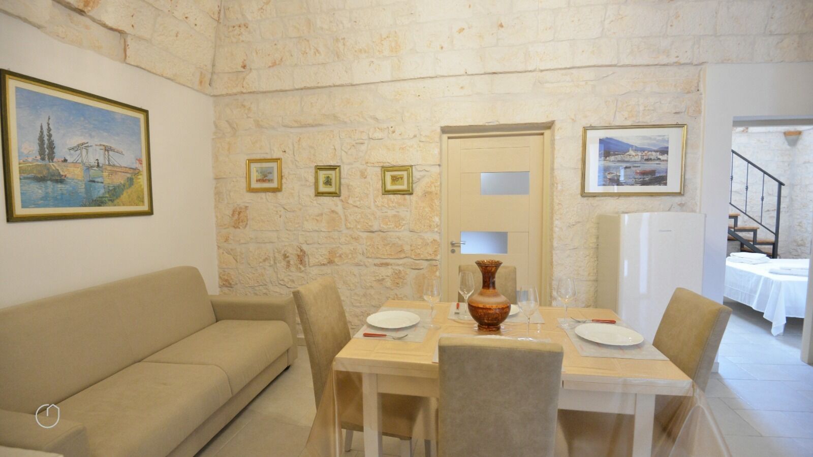 Un comedor con pared de piedra, una mesa de madera preparada para dos personas, un sofá color beige y cuadros en la pared.