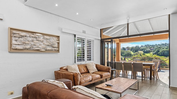A spacious living area with a brown leather sofa, a coffee table, and a dining table with chairs. There is a large window providing a view of greenery outside. - San Lameer Villa 2533 (Southbroom)
