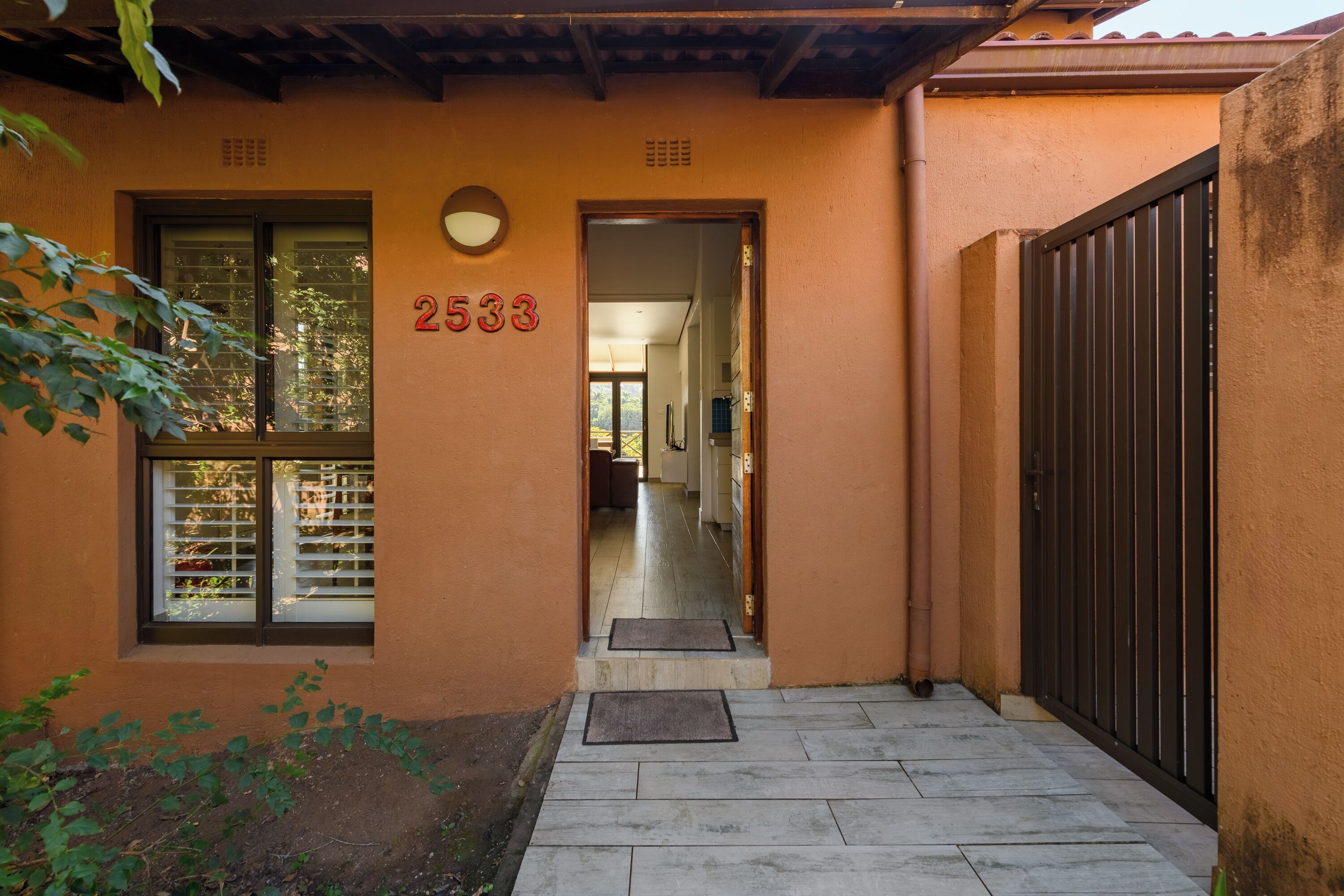 Entrada de um edifício com o número '2533' na parede, uma janela com persianas, uma porta e um caminho pavimentado que dá acesso ao interior.