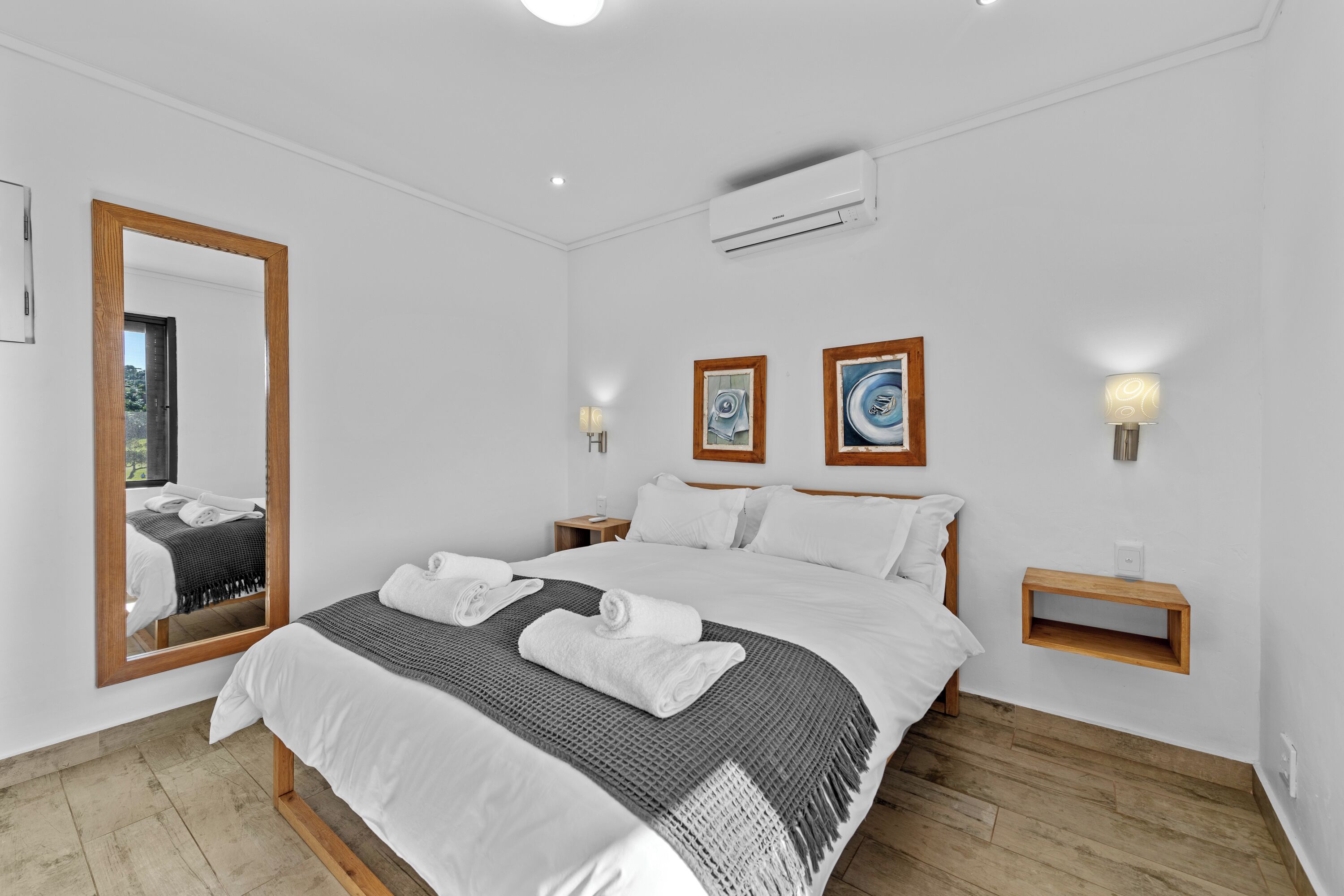 A modern hotel room with a large bed, a wooden-framed mirror, two bedside tables with wall-mounted lamps, and a small wooden shelf.
