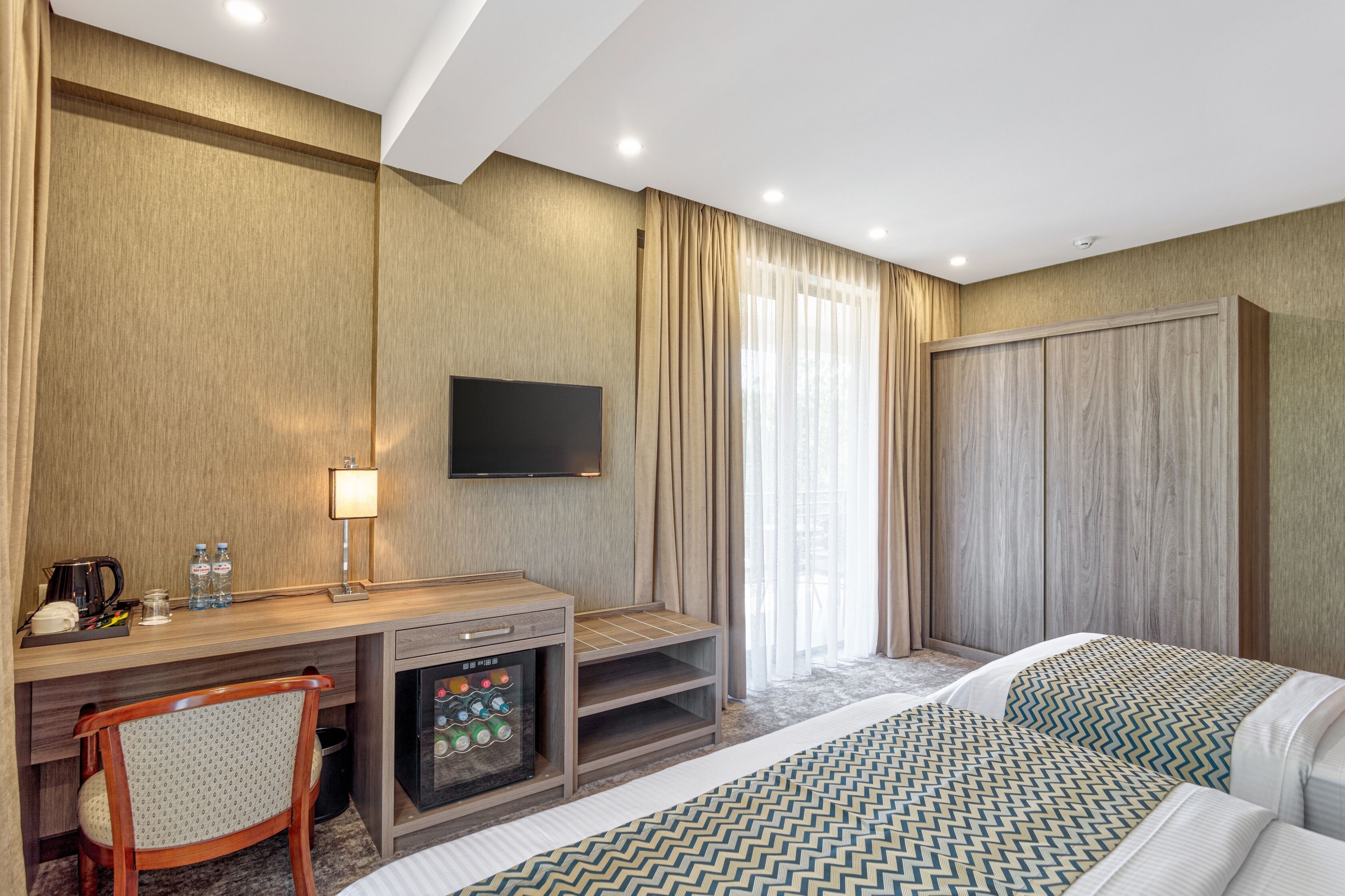 A hotel room with a bed, a desk with a TV, a minibar, and a large window with curtains.