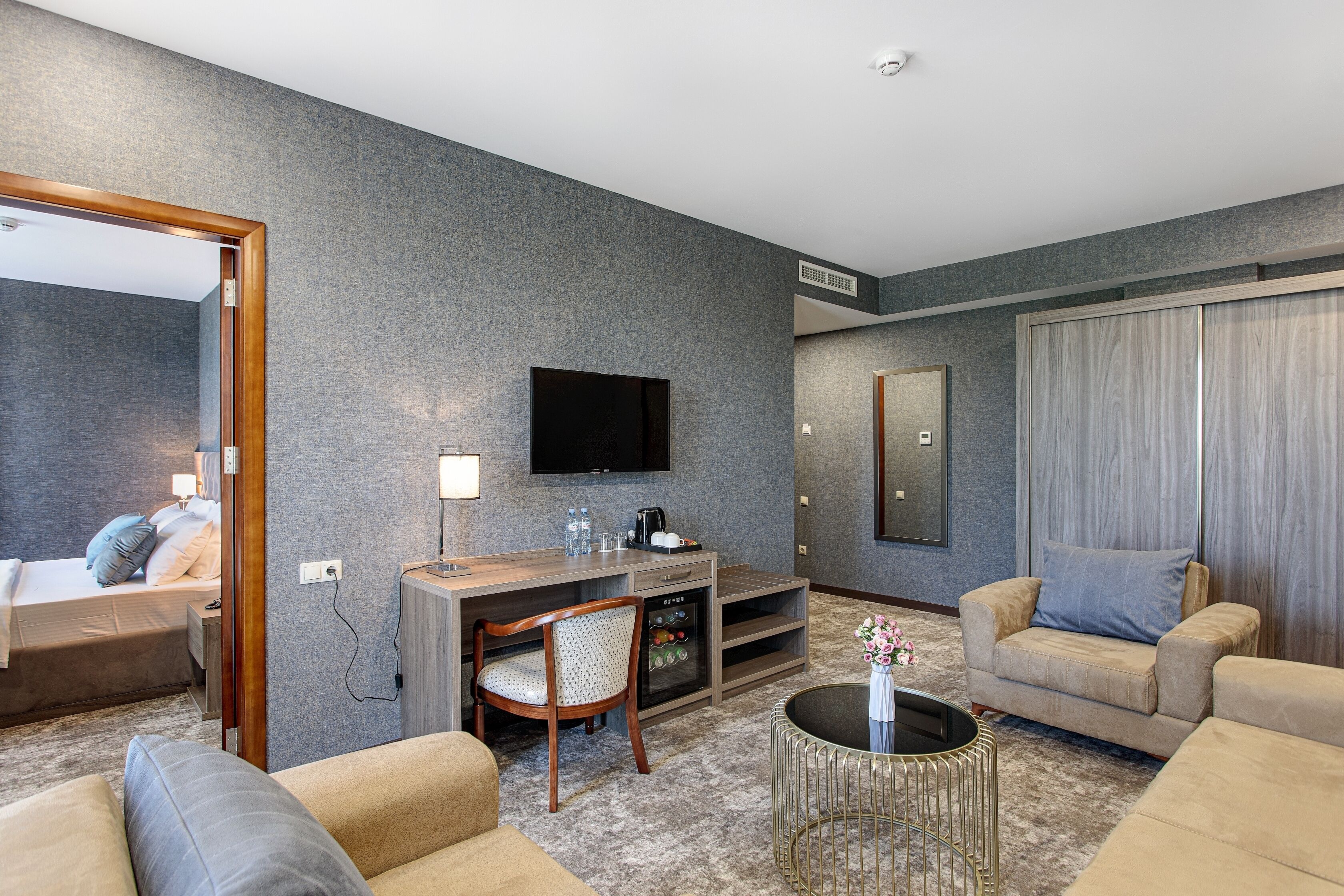 A modern hotel room with a large bed, a sofa, a coffee table, a TV, and a minibar.
