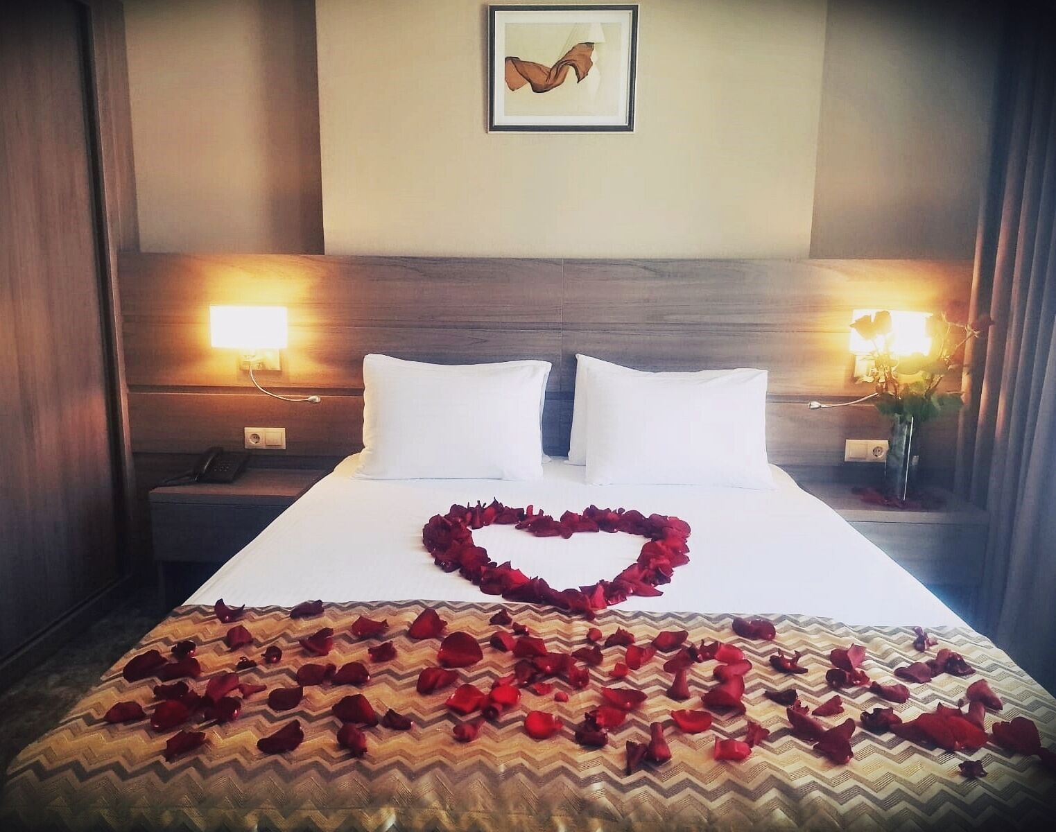 Quarto de hotel com uma cama, duas luminárias de cabeceira, uma mesa de apoio, um quadro na parede e um arranjo de pétalas de rosa em formato de coração na cama.